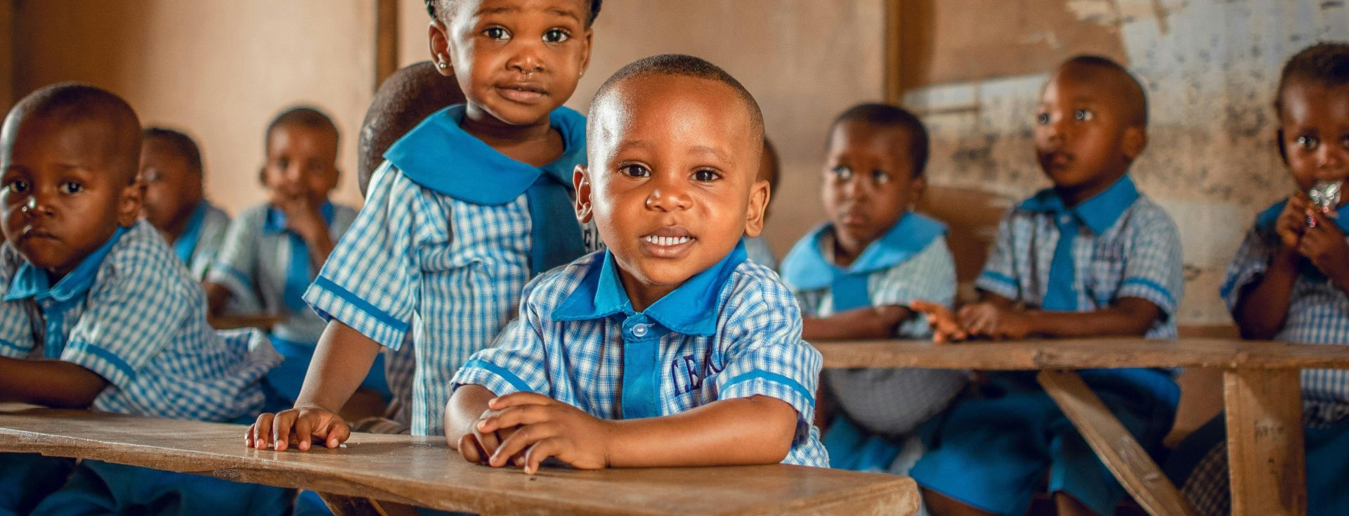 Happy children in a Nigerian classroom wearing uniforms, sitting on wooden benches.